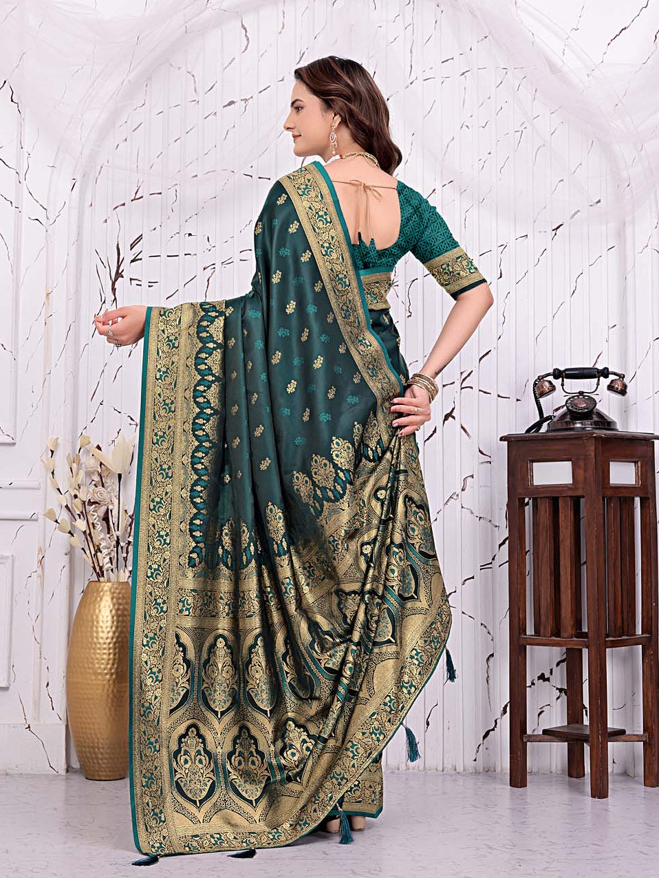 A woman models a dark teal green saree with intricate gold and beige floral patterns and a matching green blouse, standing in front of a white arched wall with decorative props.