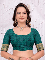 A woman modeling a teal green printed blouse with gold patterned sleeve borders, paired with gold jewelry, against a light textured wall.