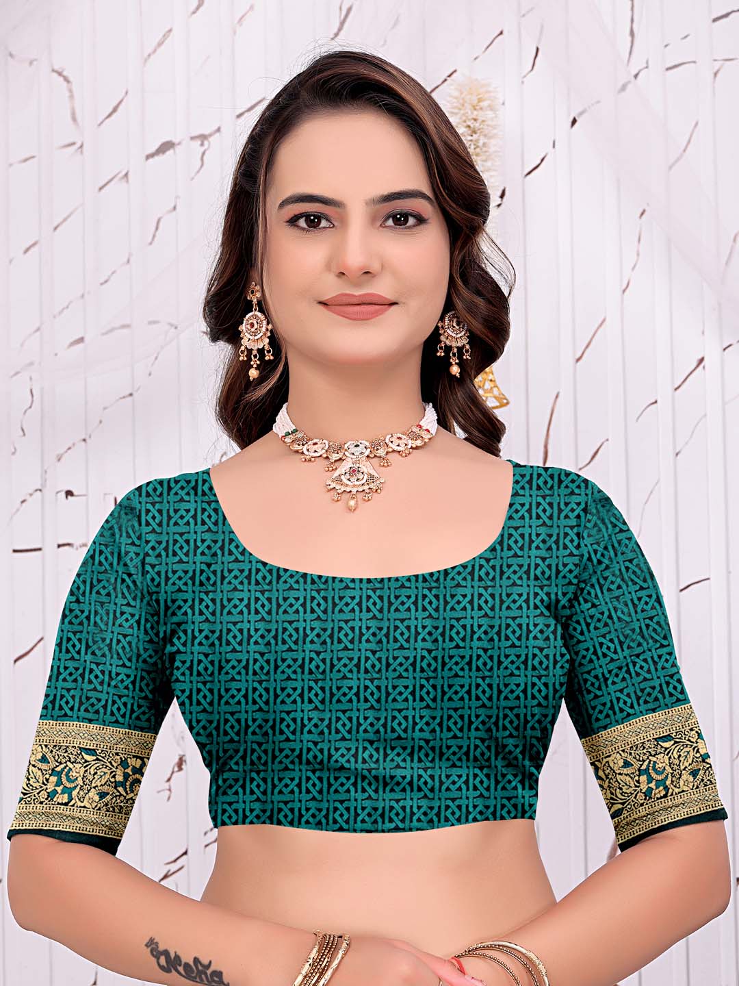 A woman modeling a teal green printed blouse with gold patterned sleeve borders, paired with gold jewelry, against a light textured wall.