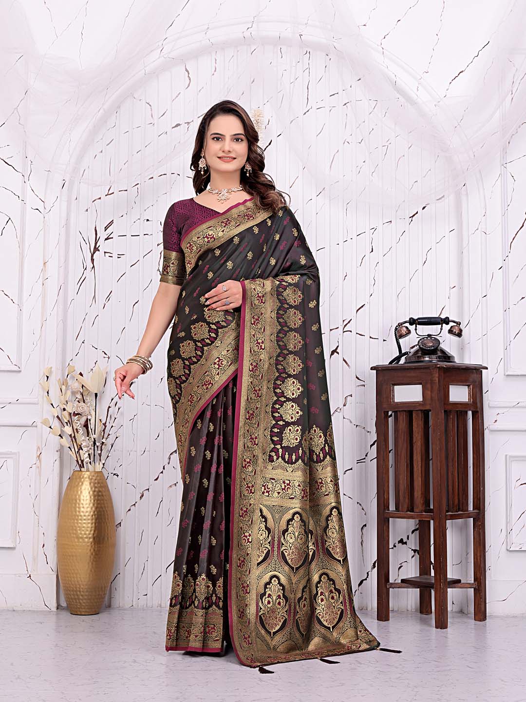 A woman models a dark brown and gold Banarasi silk saree with a deep purple blouse, standing in front of a white arched wall decorated with a gold vase and a wooden side table.
