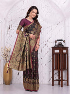 A woman models a traditional Indian saree in a decorated indoor setting with a white arched wall, a wooden table with a vintage telephone, and a gold vase. The saree has a dark purple and black base with intricate gold and red floral patterns.