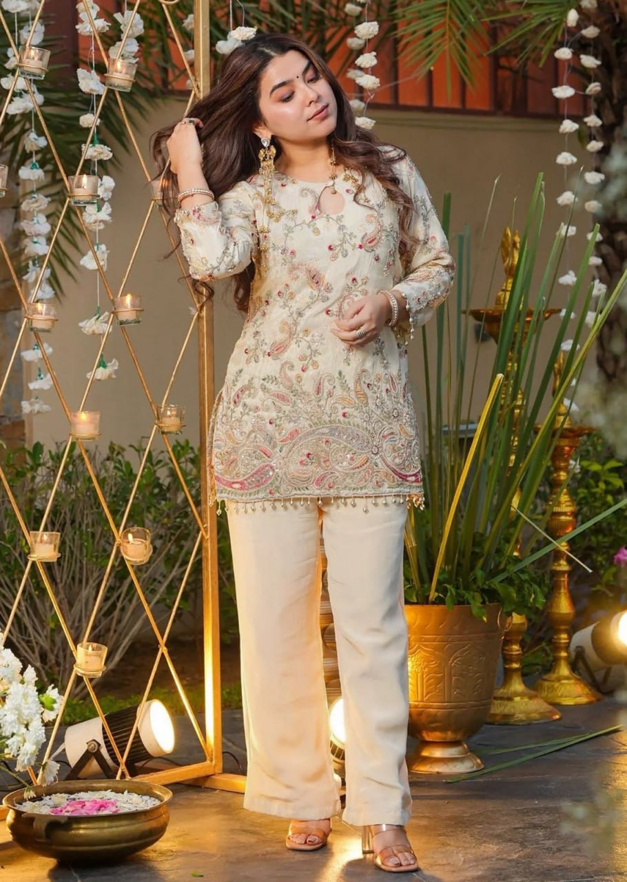 A woman models a traditional three-piece salwar kameez suit in a decorated outdoor setting. The outfit consists of a beige tunic with intricate multicolor floral and paisley embroidery, paired with matching beige wide-leg trousers and a beige collar.